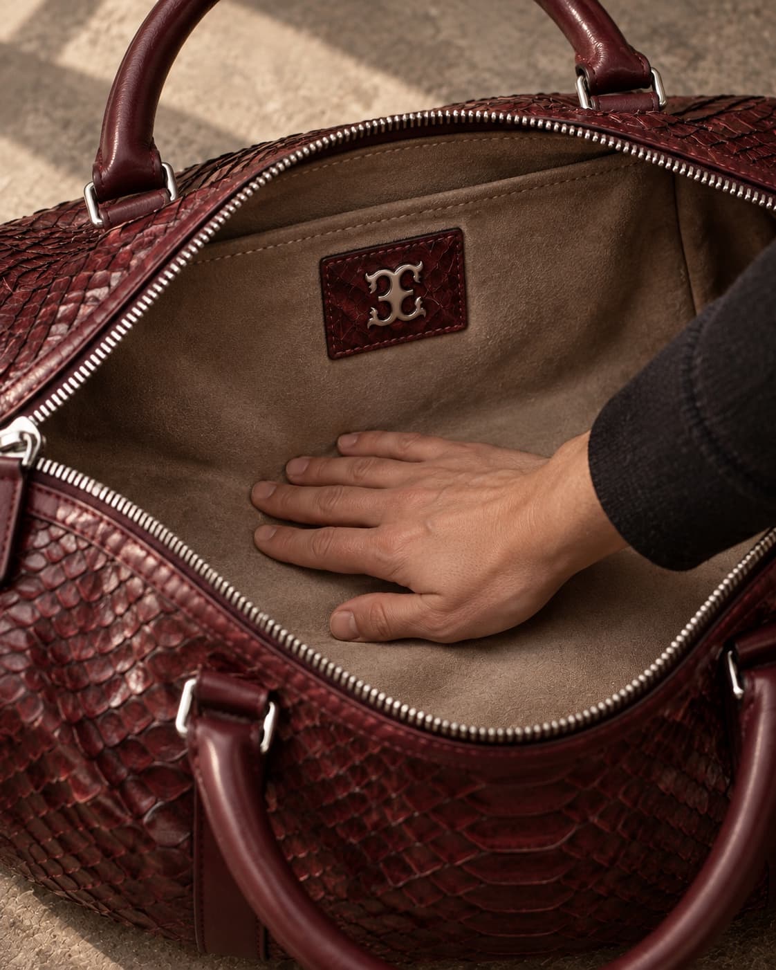 A hand smoothing the suede lining of an open python duffel, its silver seal patch on the lining wall